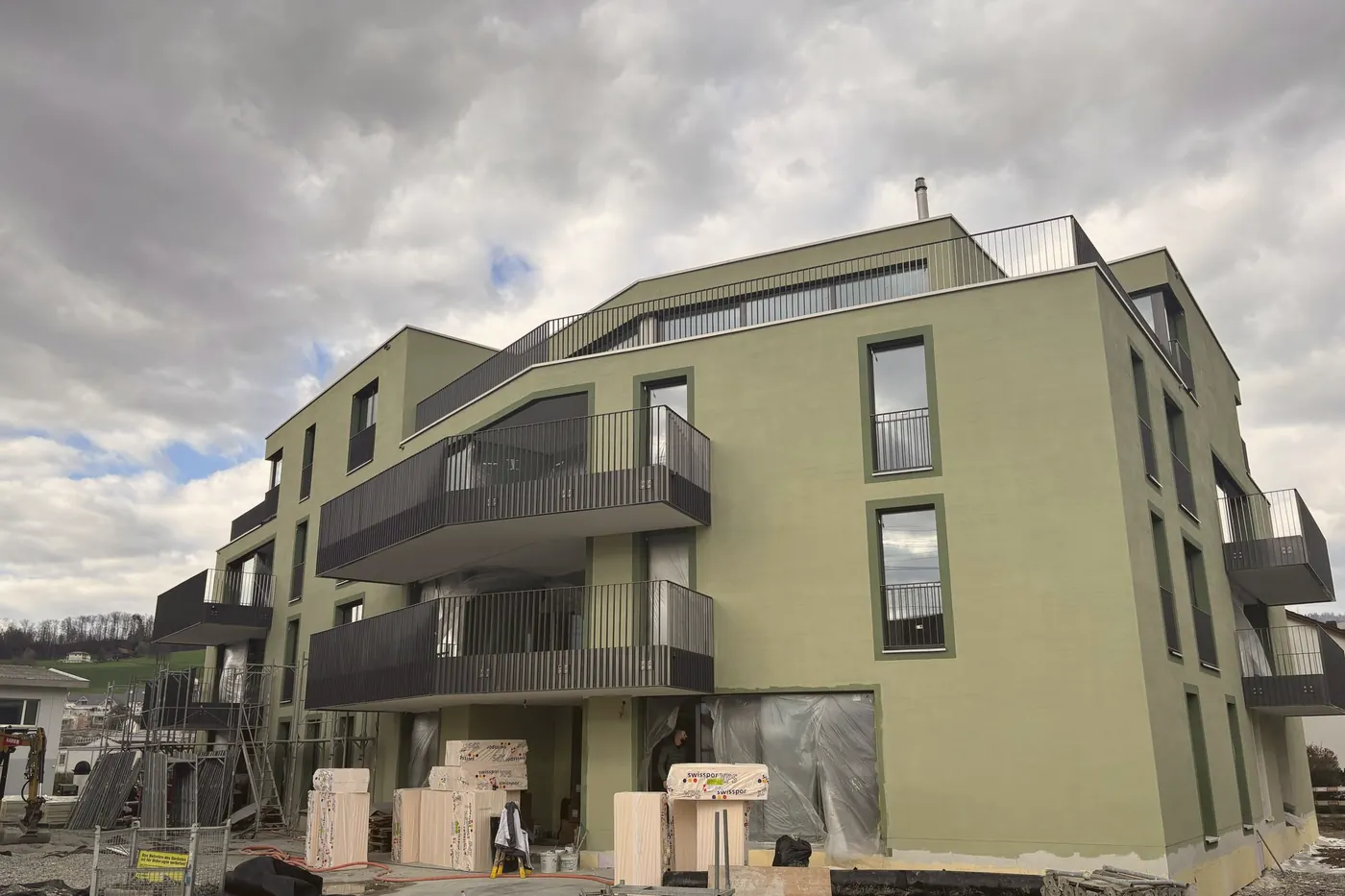 Neubau mit grüner Fassade und dunklen Balkonen, davor Baumaterial