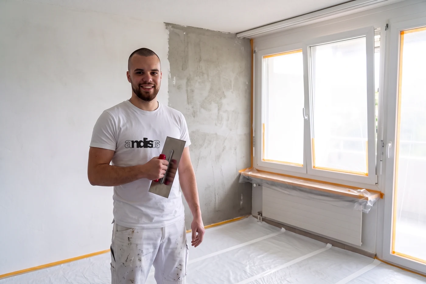 Gipser im Andisa-Shirt mit der Glättekelle vor einer halb verputzten Wand