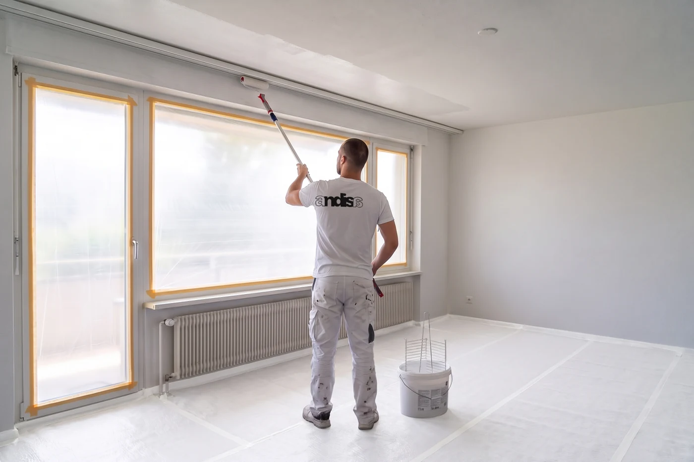 Maler im Andisa-Shirt streicht die Decke mit der Teleskoprolle entlang der Fensterwand