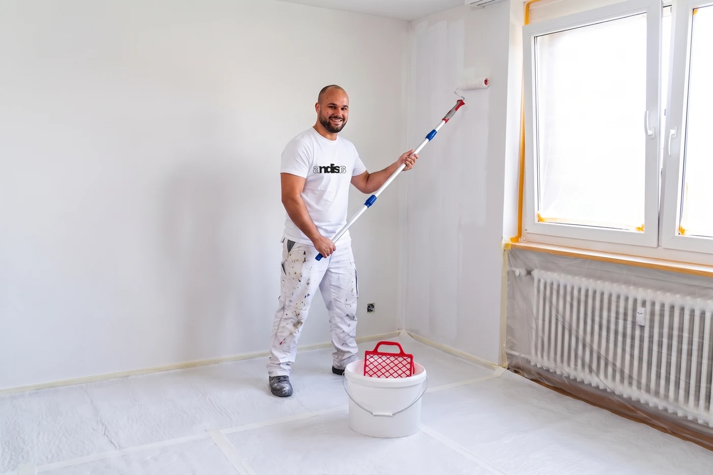 Maler im Andisa-Shirt rollt Wandfarbe mit der Teleskopstange, Boden und Fenster abgedeckt